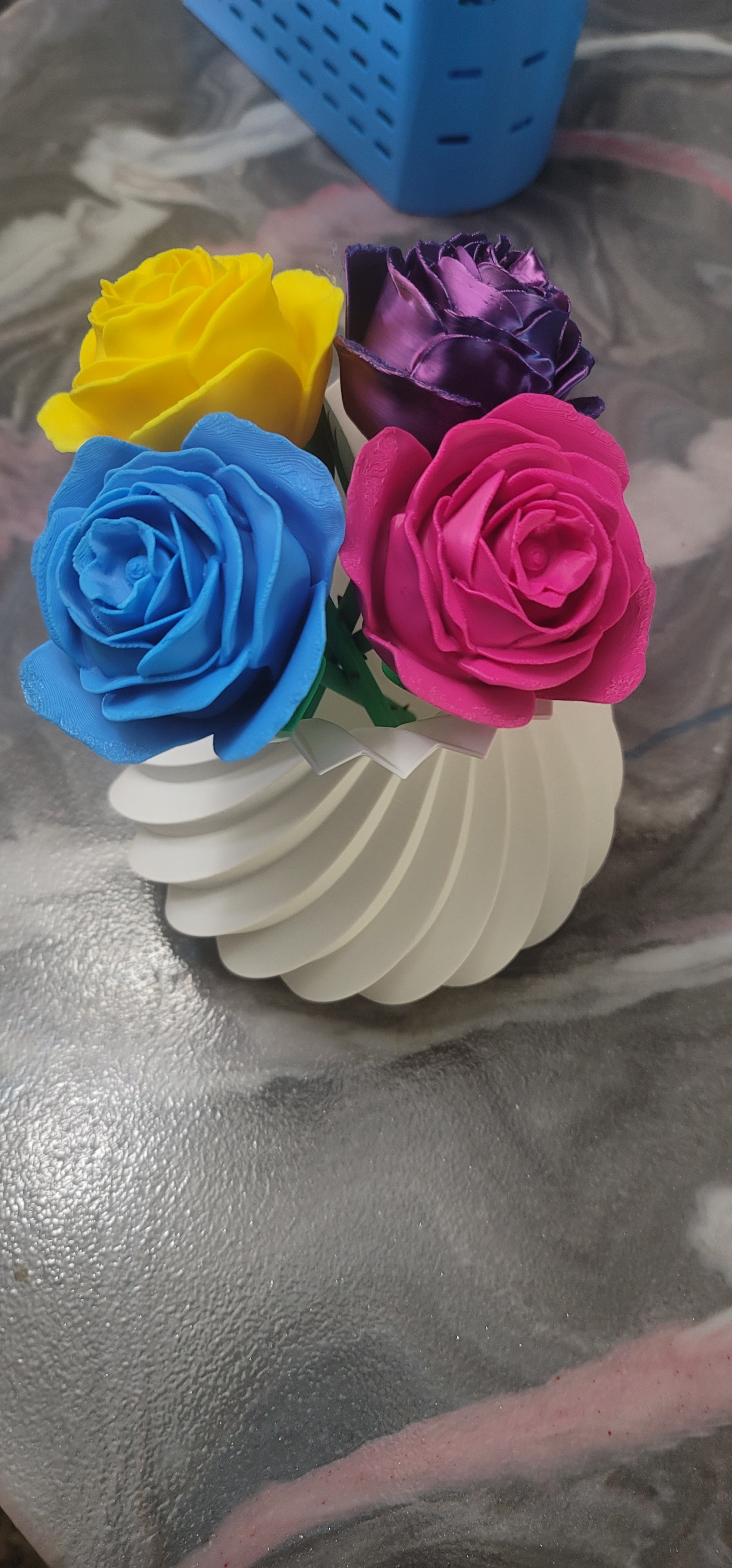 Four colorful 3D printed roses in yellow, blue, purple, and pink arranged in a white spiral vase, front view