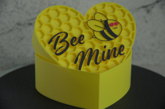 Yellow heart-shaped box with a bee design and 'Bee Mine' text on a dark surface.