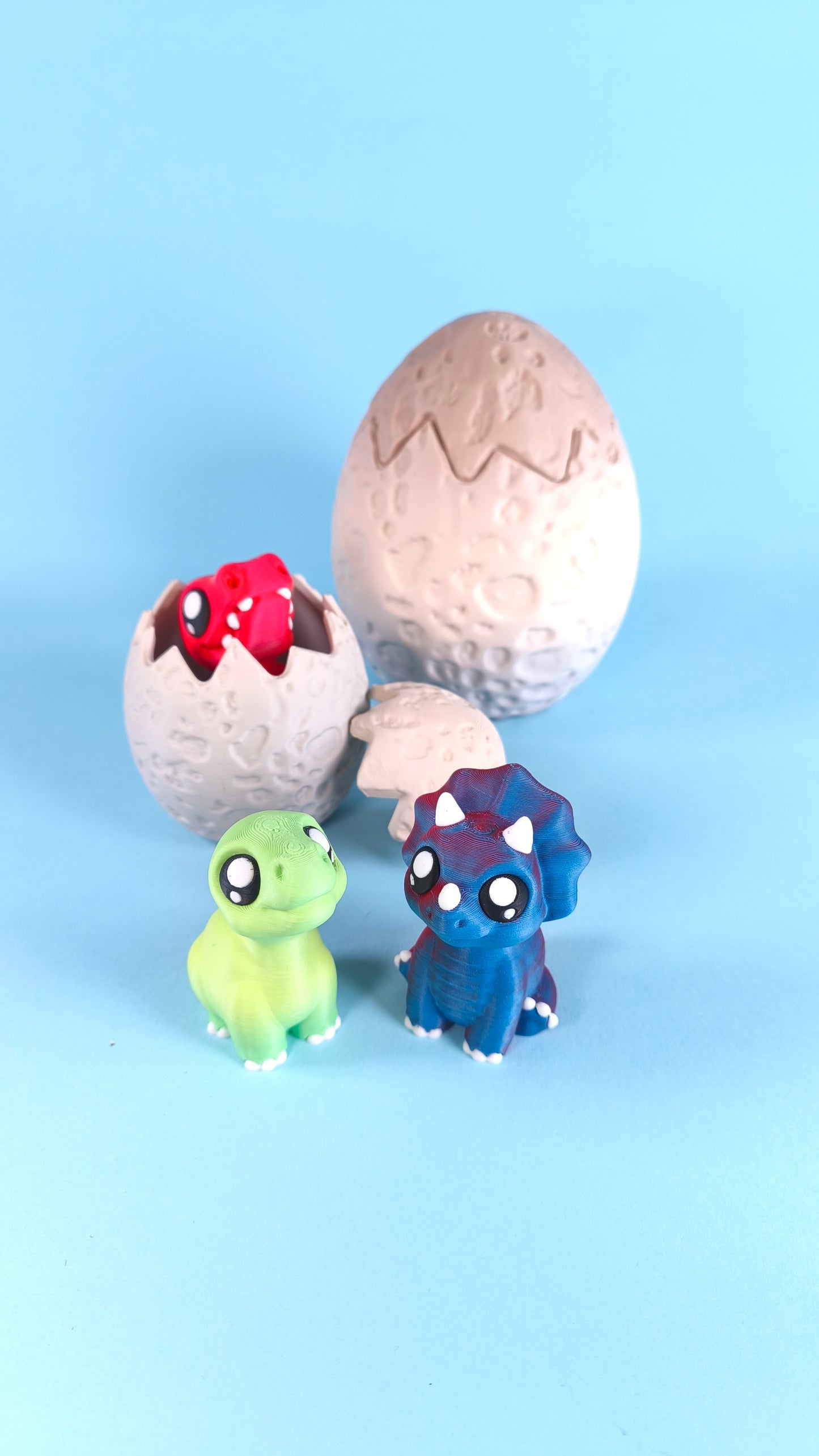 3D printed baby dinosaur finger puppets — red T-rex and green dino with blue triceratops peeking from cracked egg on a blue background