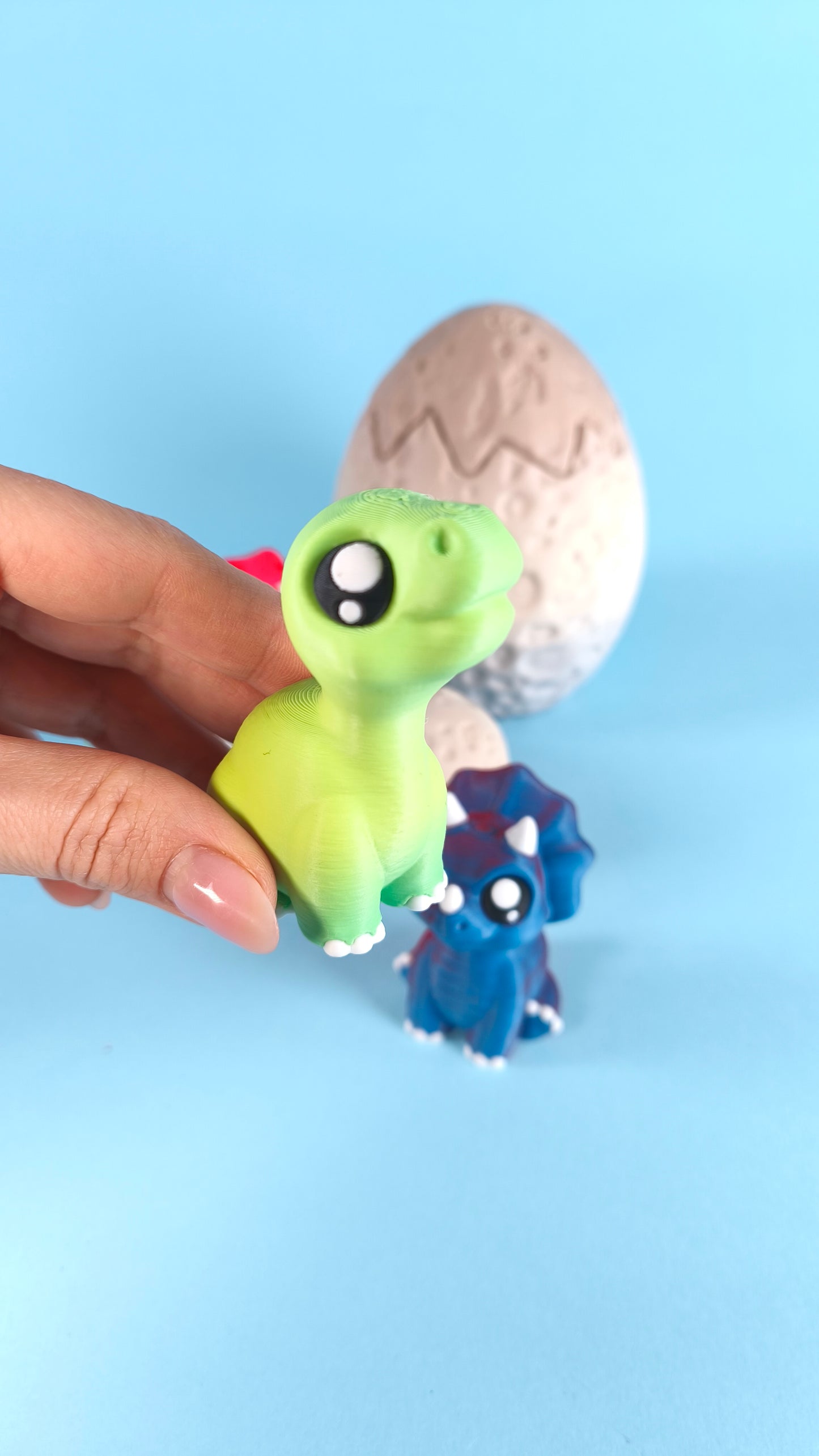 Hand holding a 3D printed green baby T-rex finger puppet with the cracked dino egg behind it and blue triceratops in the background