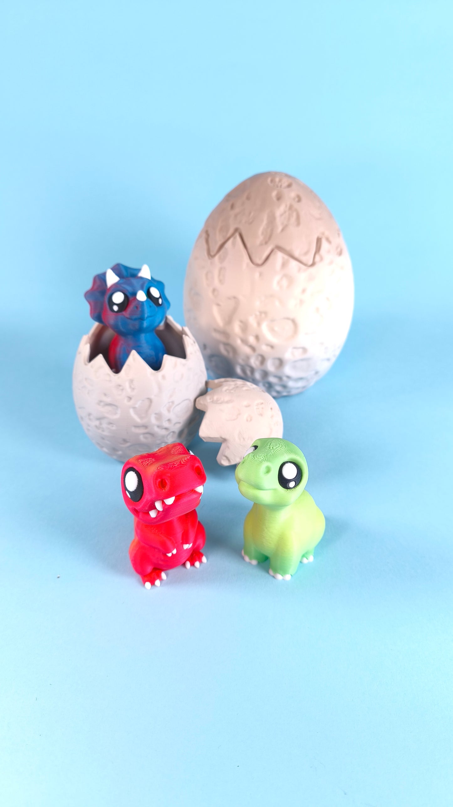 3D printed baby dinosaur finger puppets with hatching egg — red T-rex and green dino figures with blue triceratops peeking out of cracked egg