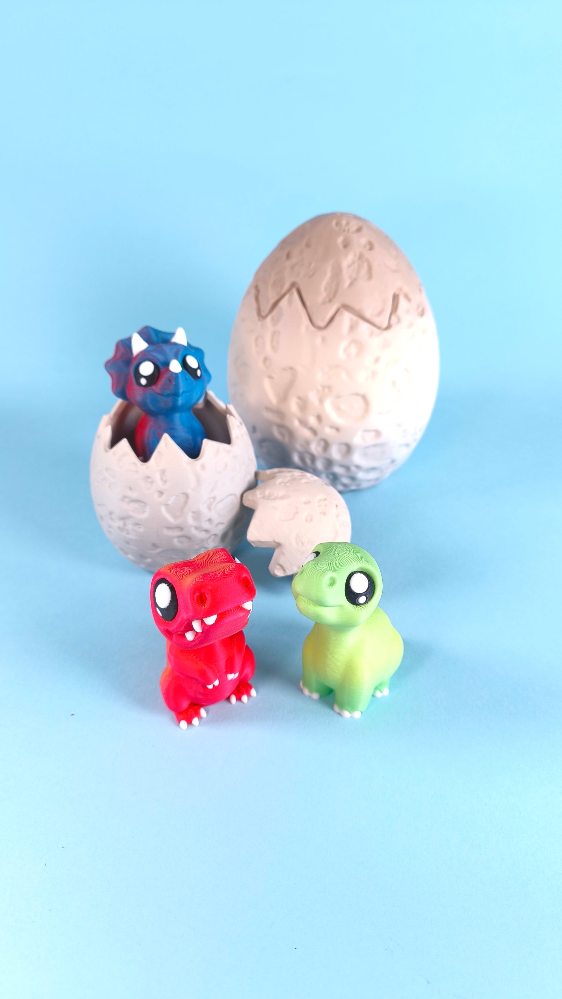 3D printed baby dinosaur finger puppets with hatching egg — red T-rex and green dino figures with blue triceratops peeking out of cracked egg