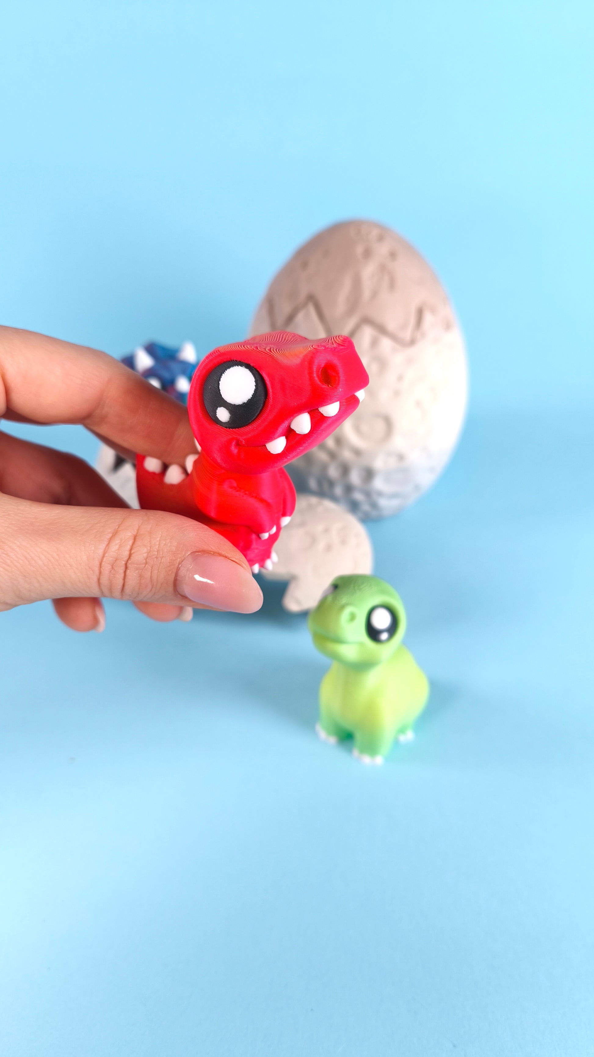 3D printed Finger Dinosaur Pack — green T-rex, blue triceratops, and red T-rex baby dino figures alongside cracked dino egg shells
