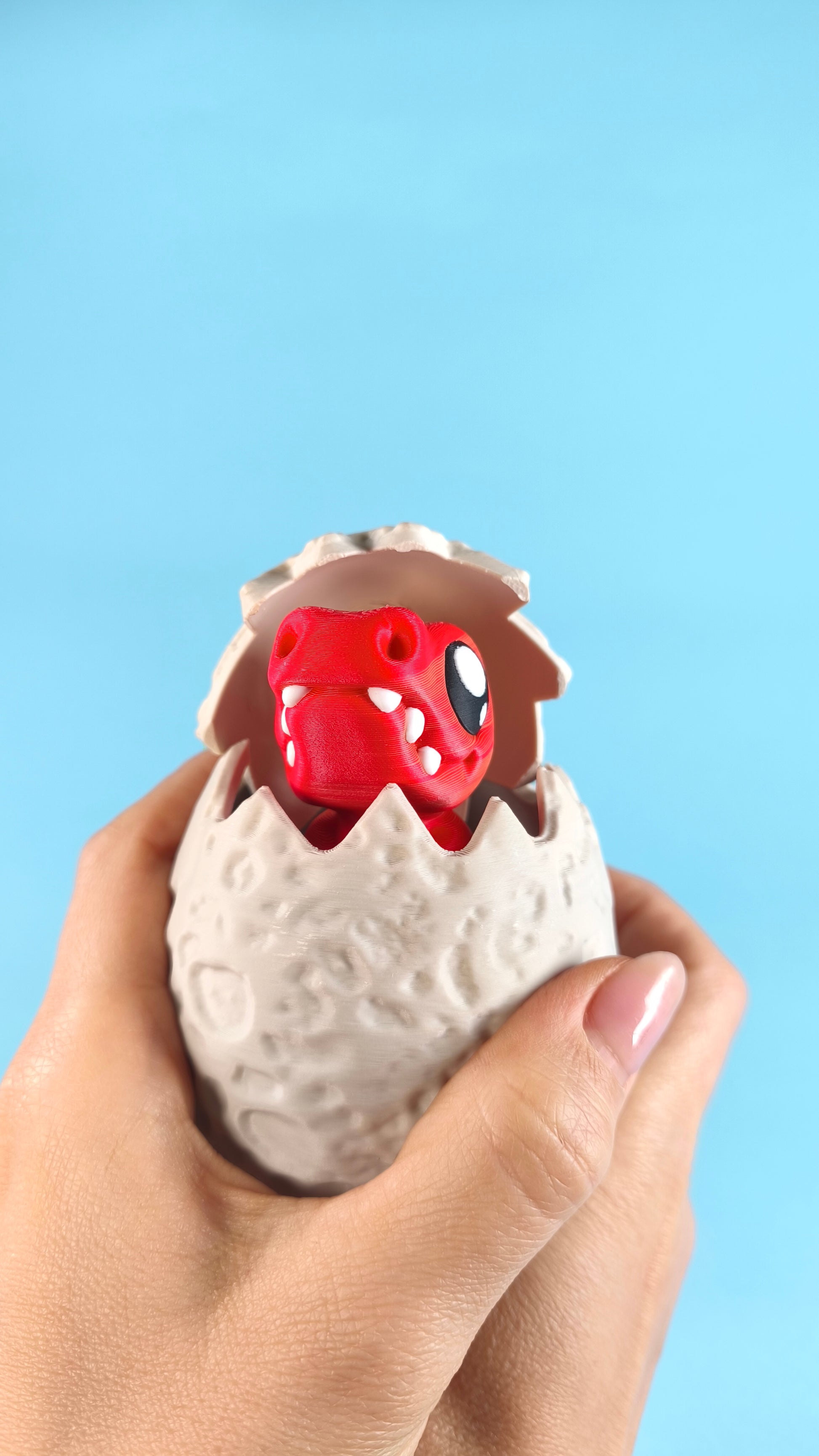 Hand holding a 3D printed red baby T-rex finger puppet close-up, with green and blue triceratops dinos visible in the background