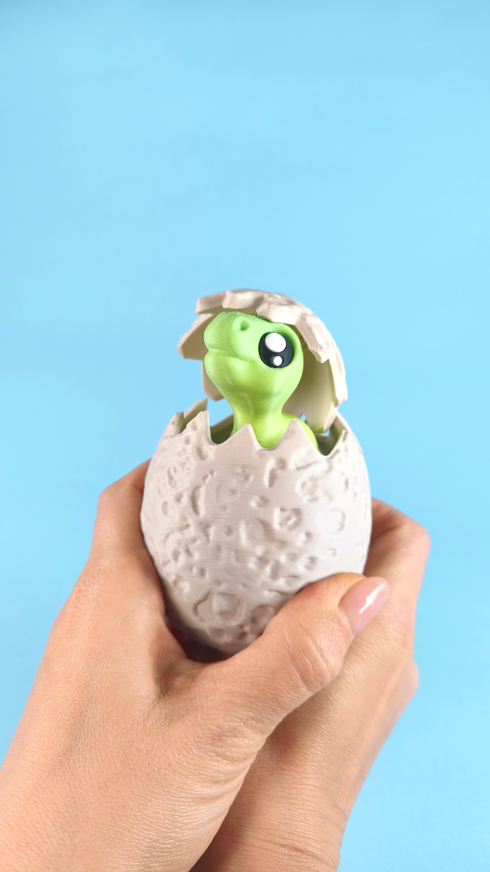3D printed green baby T-rex finger puppet peeking out of a cracked dino egg held in two hands on a blue background