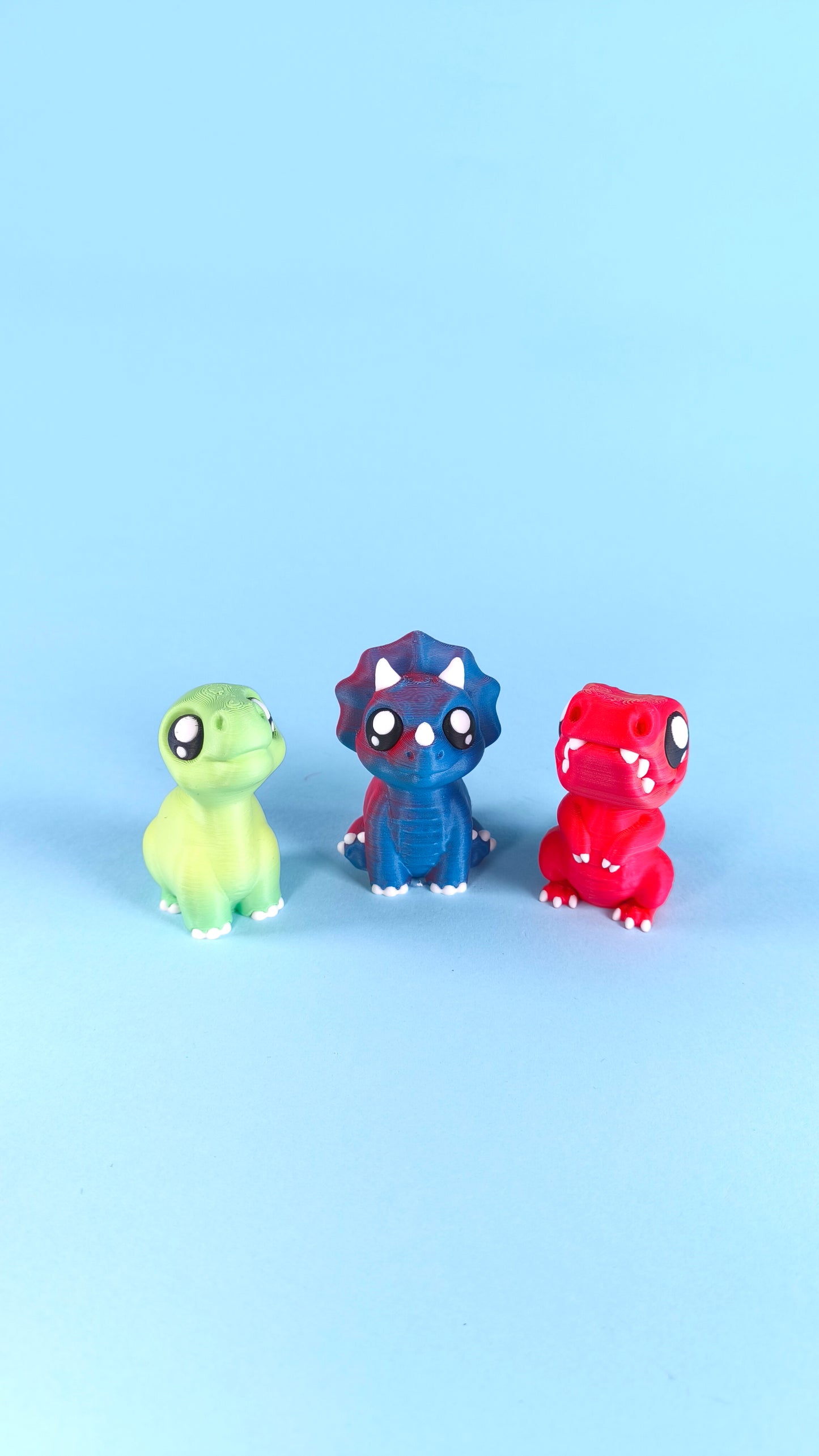 Three 3D printed baby dinosaur finger puppets — green T-rex, blue triceratops, and red T-rex — standing on a light blue background