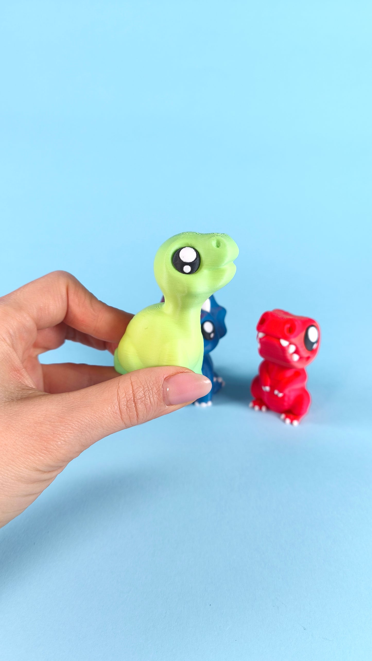 Hand holding a 3D printed green baby T-rex finger puppet to show size scale, with blue triceratops and red T-rex dinos in the background