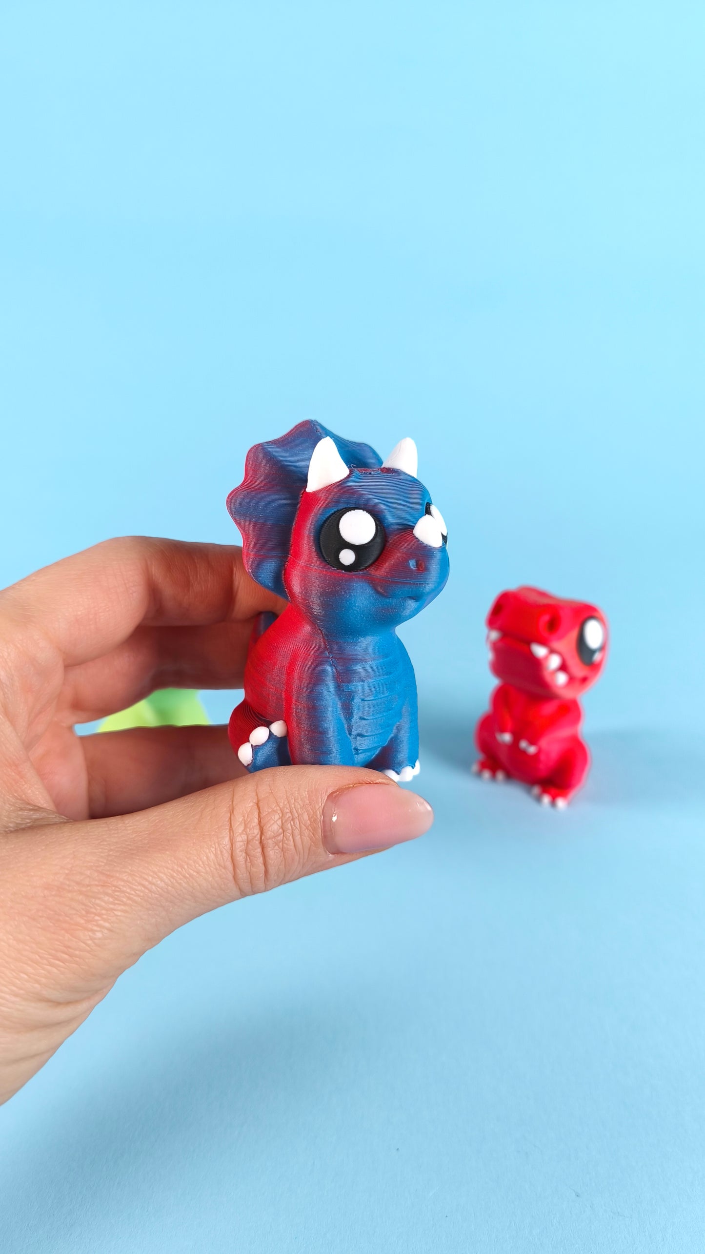 Hand holding a 3D printed blue baby triceratops finger puppet close-up, with red T-rex and green dino visible in the background