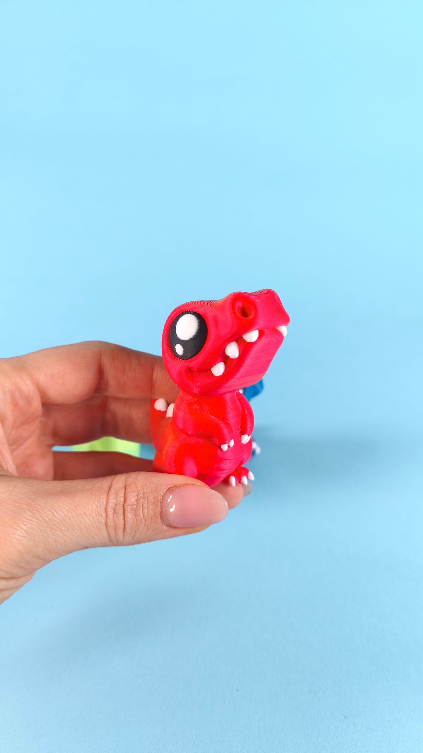 3D printed green baby T-rex finger puppet peeking out of a cracked dino egg shell held in hands on a blue background