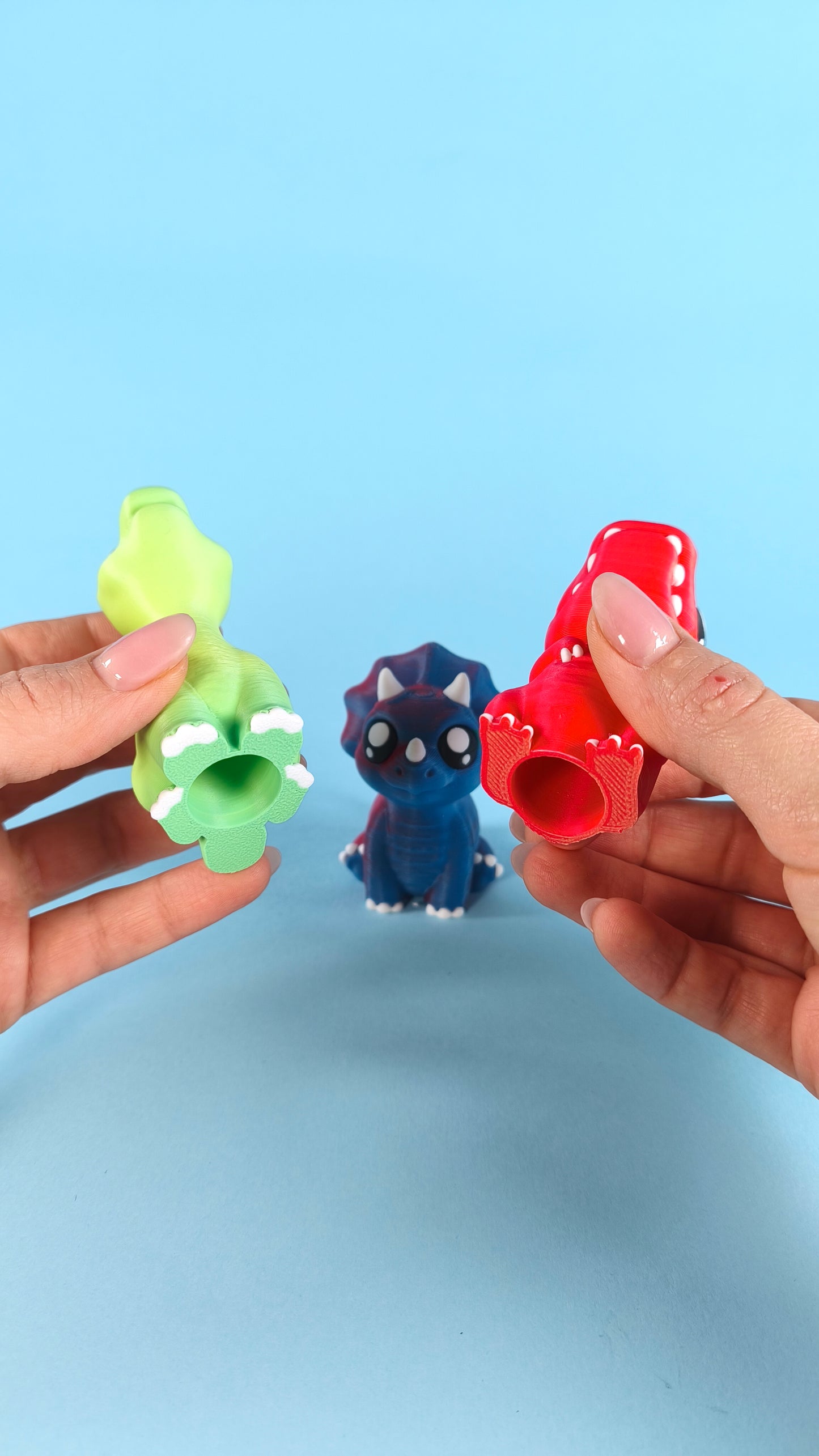 Hand holding a 3D printed red baby T-rex finger puppet showing cute face detail, with green and blue dinos in background