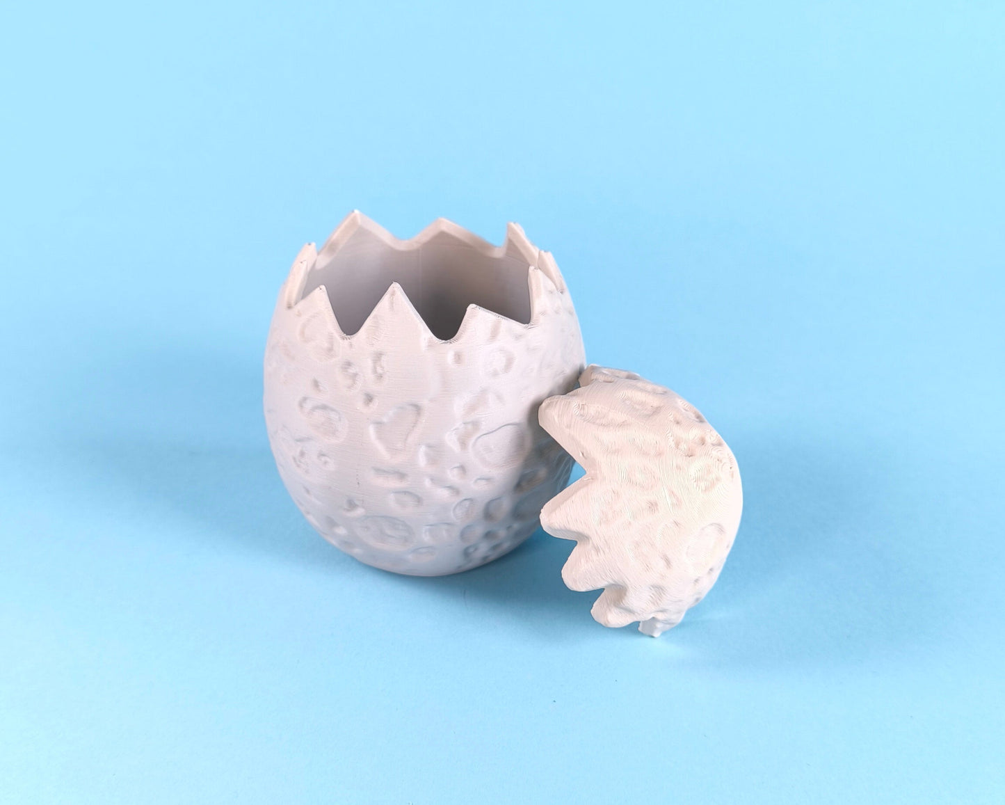 3D printed dino egg shells open and closed — showing hollow interior where finger puppet dinosaurs nest, on a light blue background